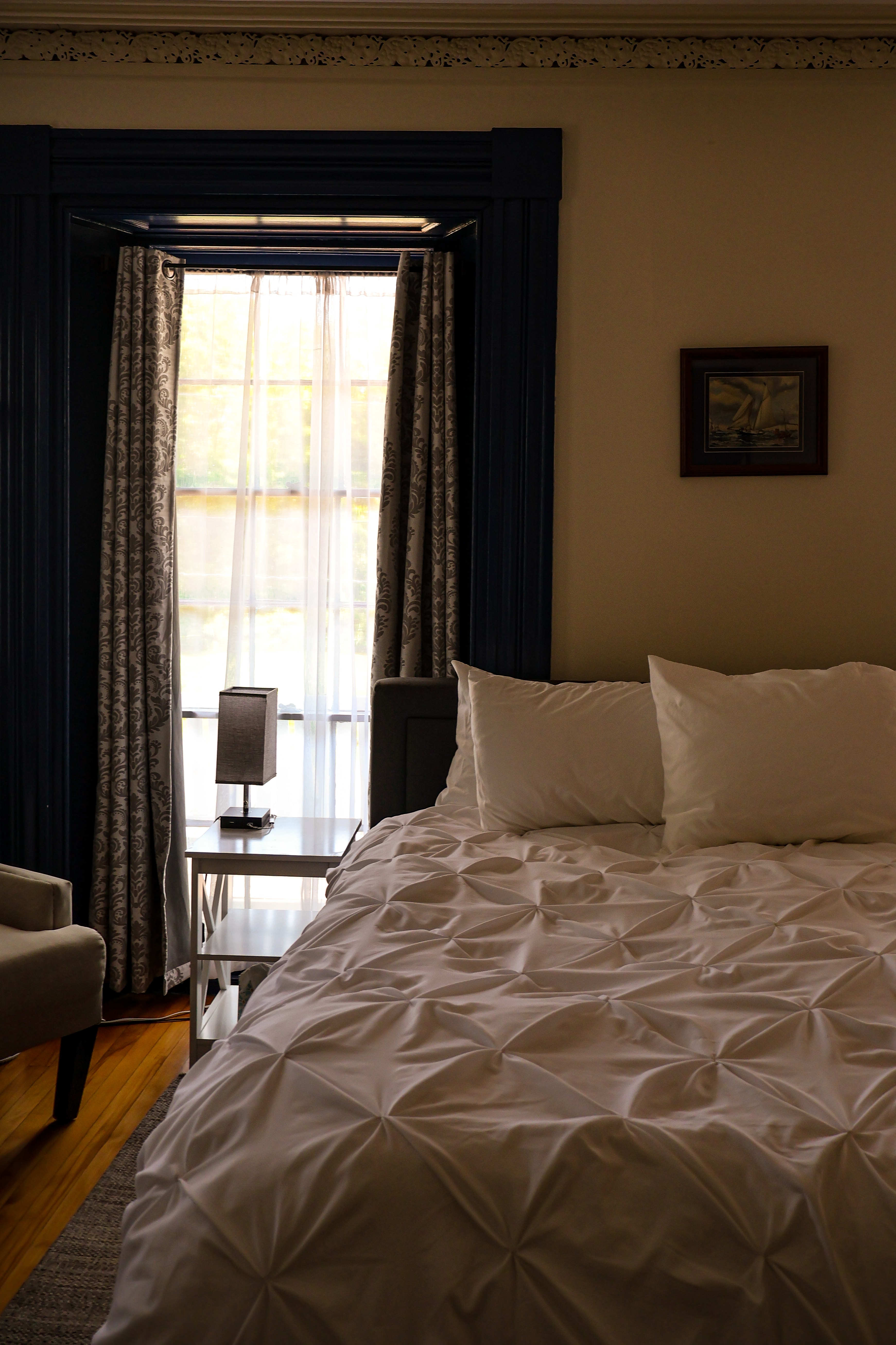 A cozy bedroom featuring a bed with a patterned white duvet, a bedside lamp, and a window with sheer curtains.