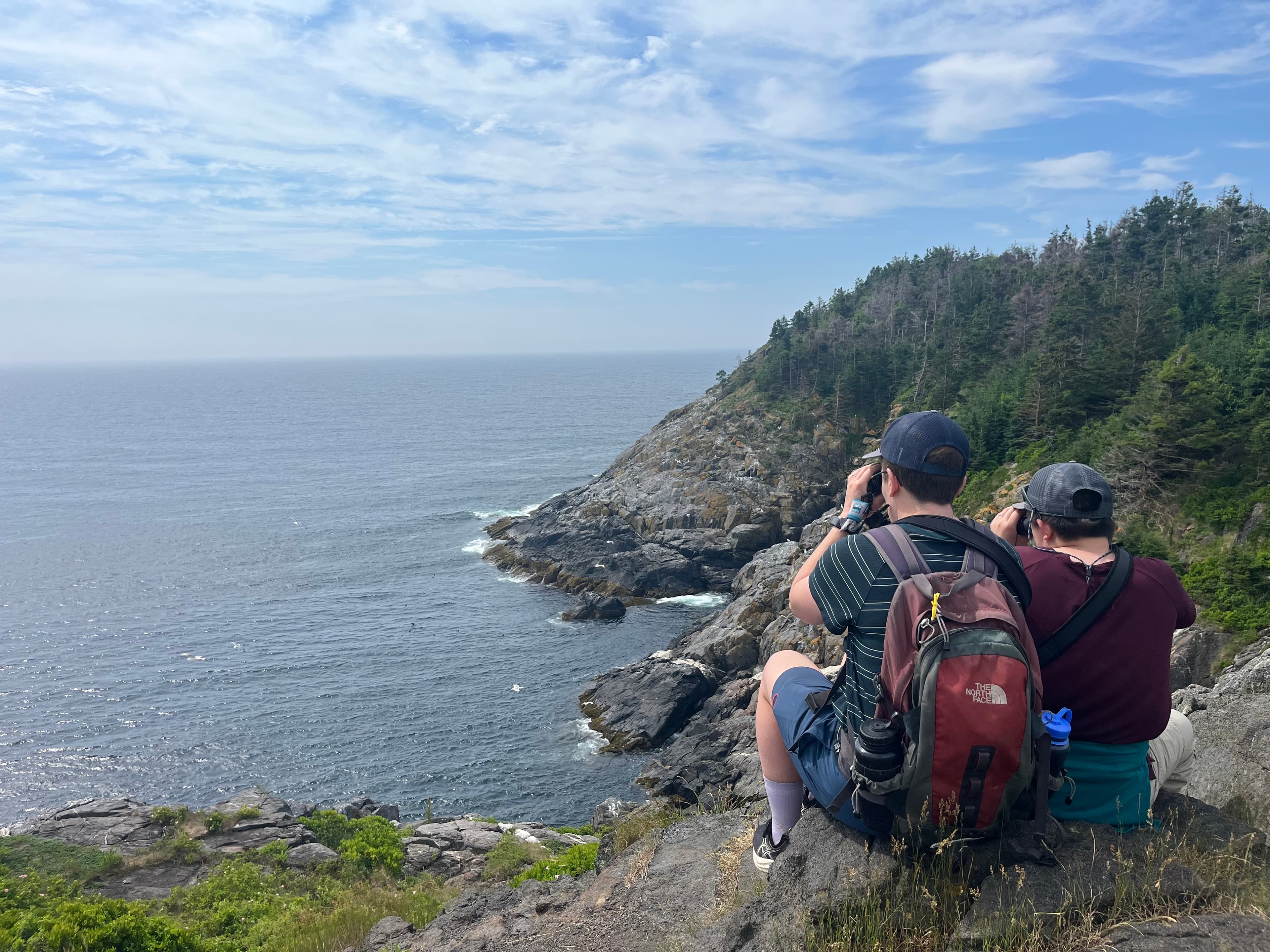 Two people sit on a rocky coastline, photographing the ocean and distant shoreline under a cloudy sky.