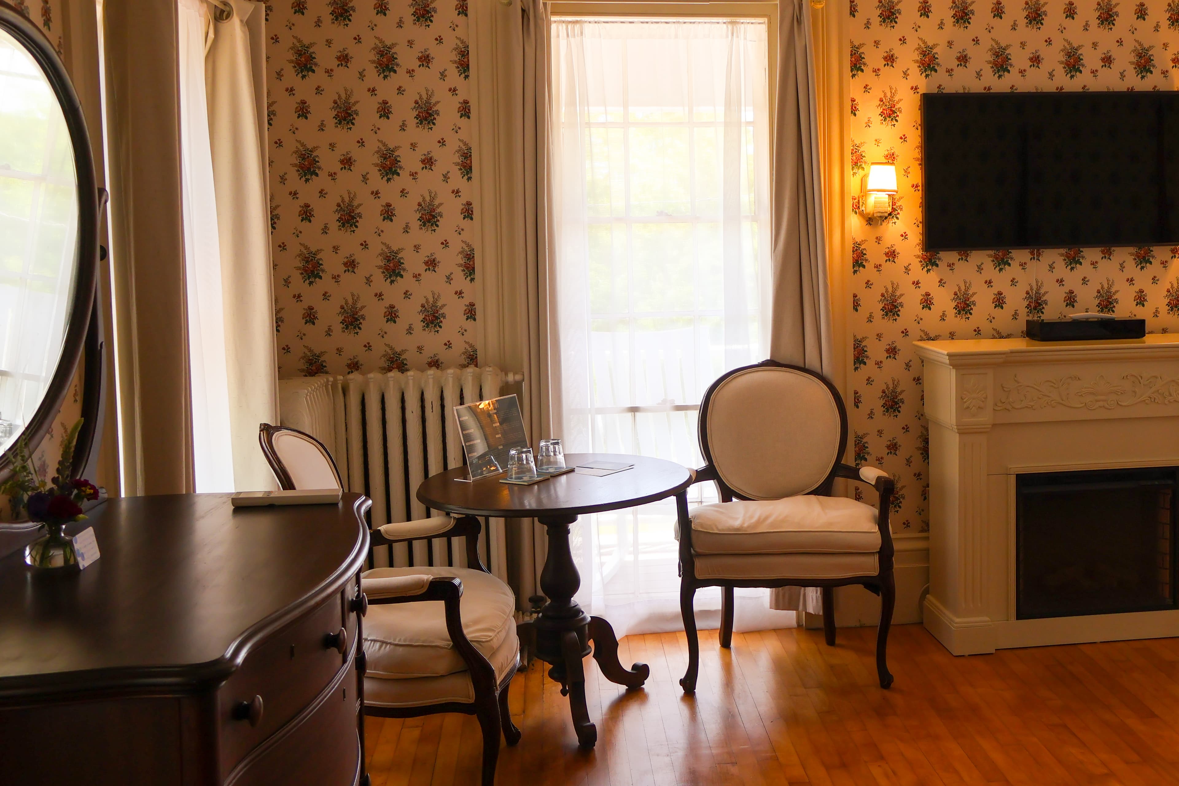 A cozy room featuring a floral wallpaper, elegant chairs, a round table, and a decorative fireplace.