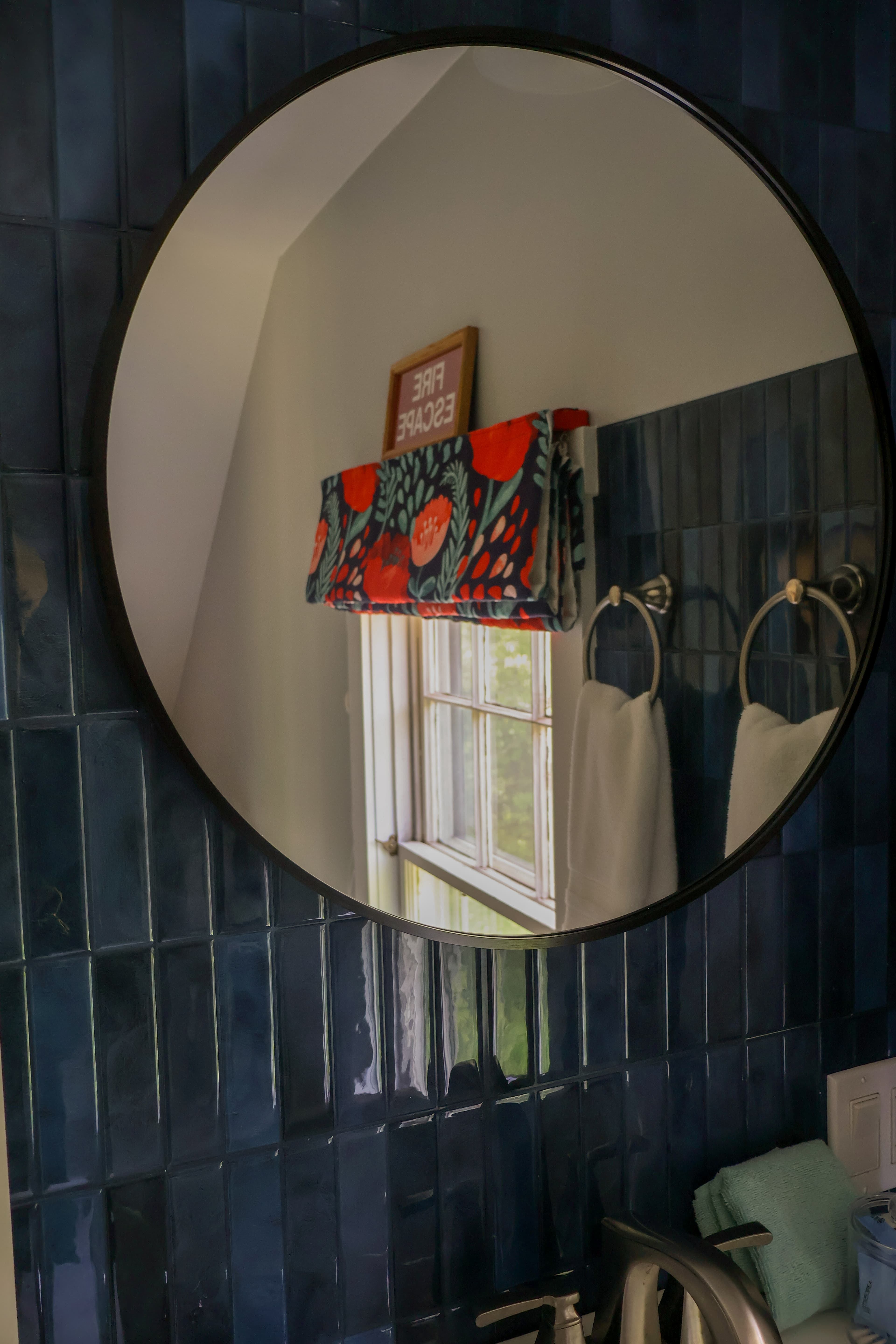 A round mirror reflects a window and patterned curtain in a bathroom with blue tiled walls.