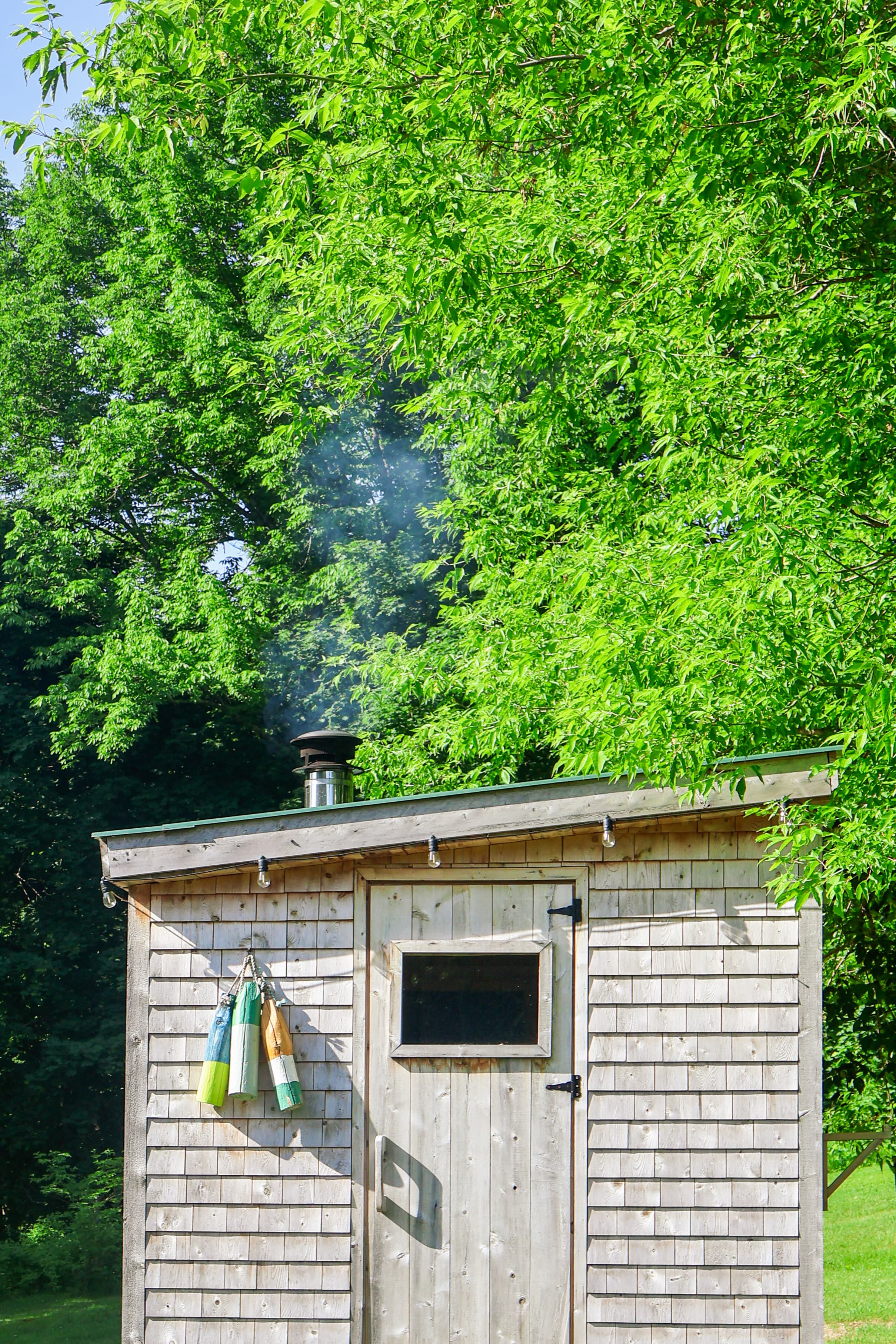 A wooden shed with colorful buoys hanging on the side and smoke rising from the chimney, surrounded by lush green trees.