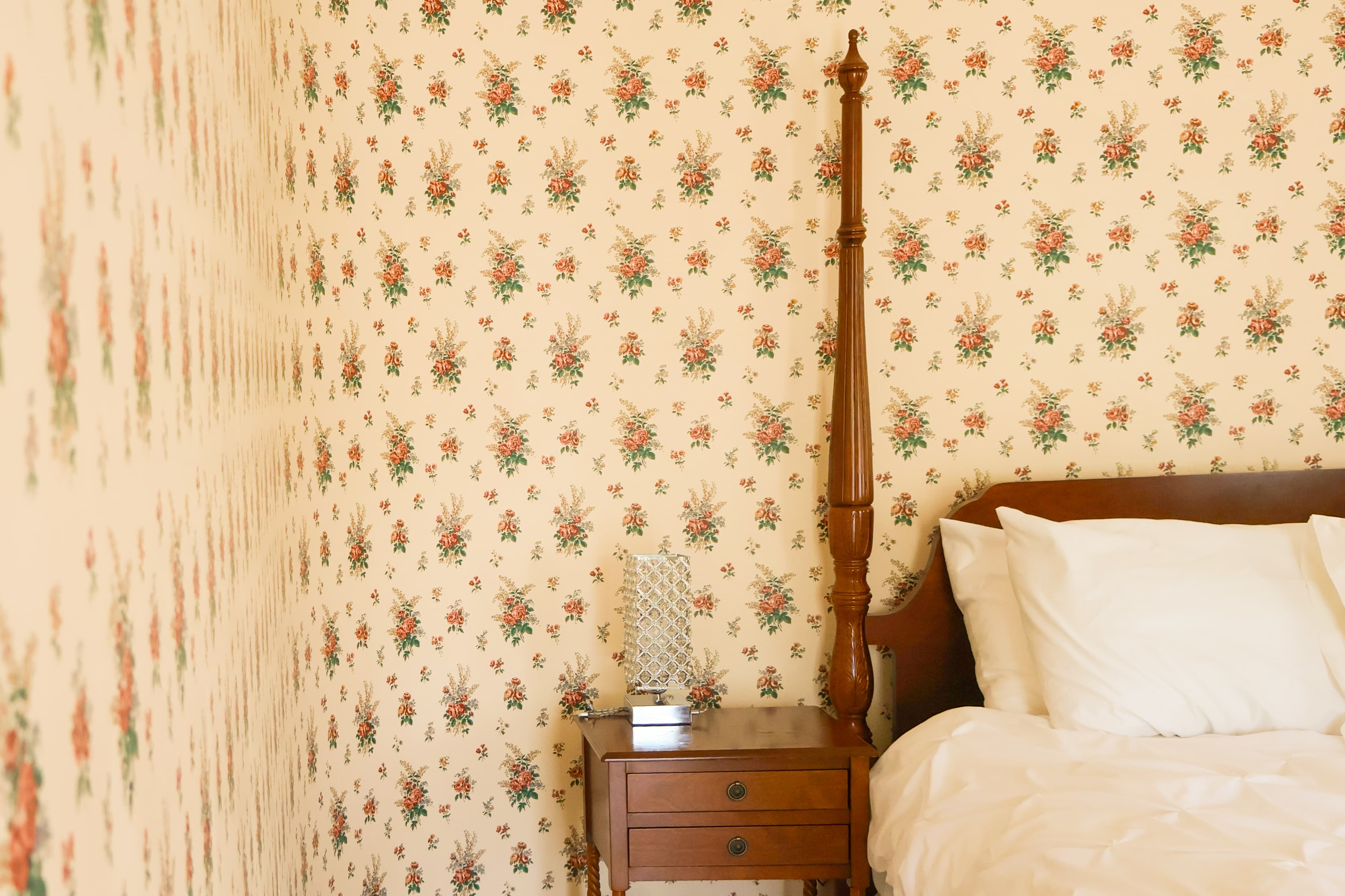 A cozy bedroom featuring floral wallpaper, a wooden bed with white bedding, and a nightstand with a decorative item.