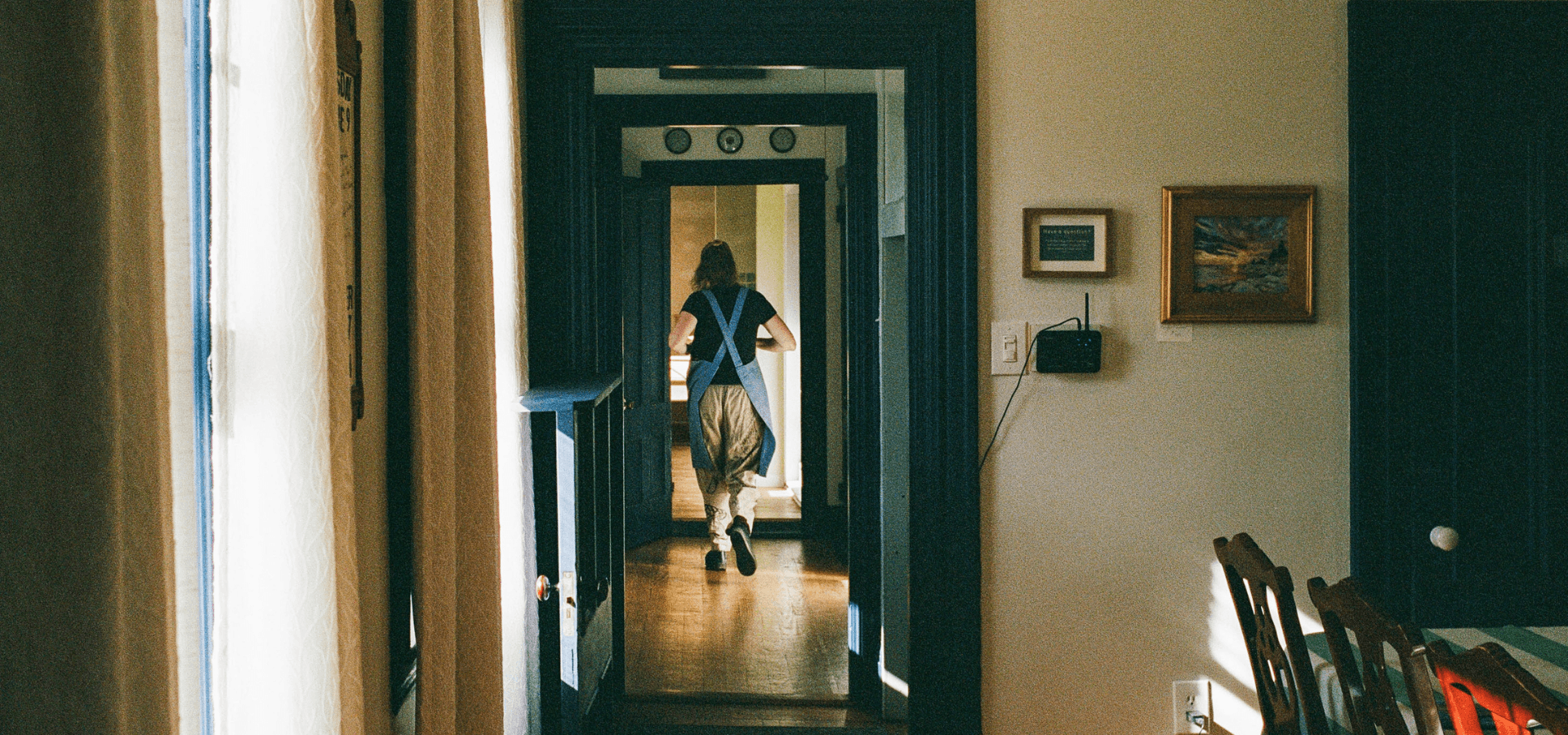 A person in overalls walks down a hallway in a warmly lit home.