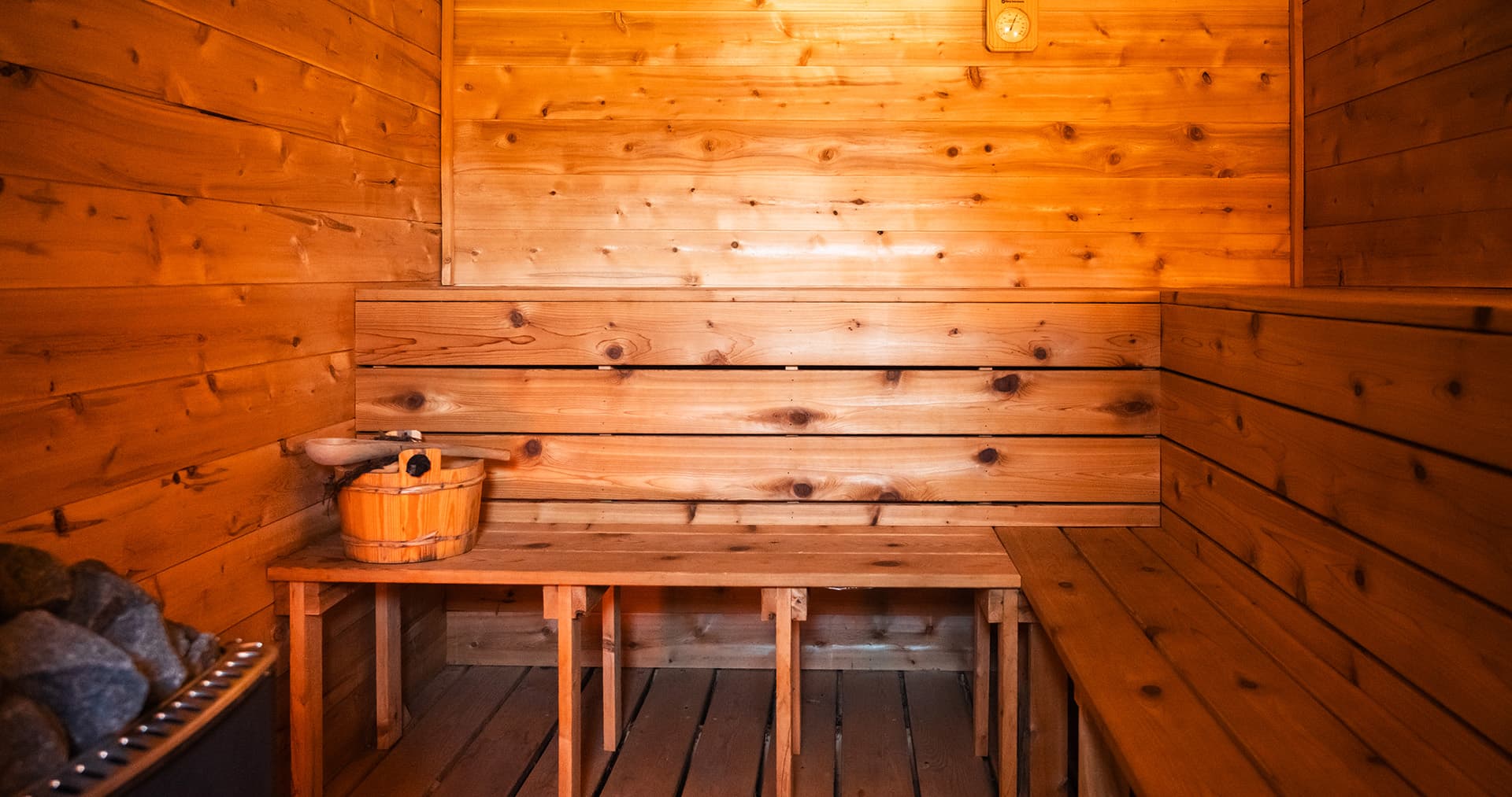 A wooden sauna interior featuring benches, a heater with stones, and a wooden bucket.