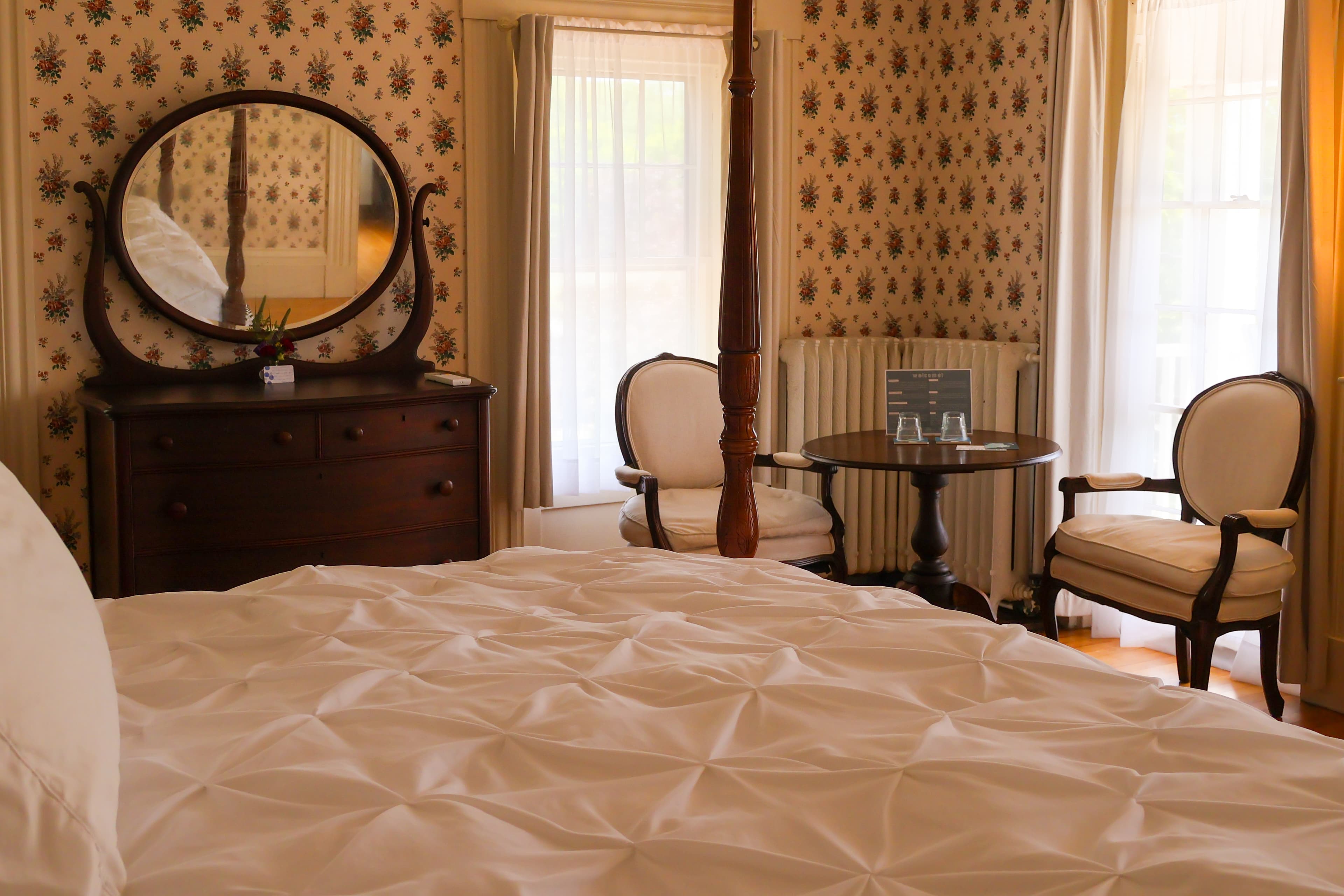 A cozy bedroom featuring a patterned wallpaper, a four-poster bed, a vintage dresser, and a seating area by a window.