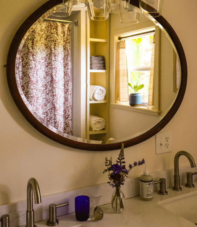 A round mirror reflects a bathroom sink area with a bouquet of flowers, artistic glassware, and a glimpse of a shower curtain and window.