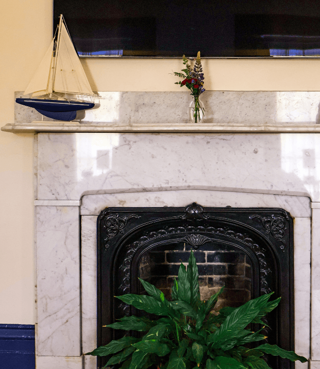 A marble fireplace mantel featuring a decorative sailboat, a small floral arrangement, and a leafy plant.