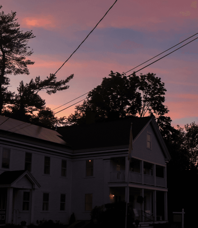 A white house is silhouetted against a colorful sunset with trees and power lines in the background.